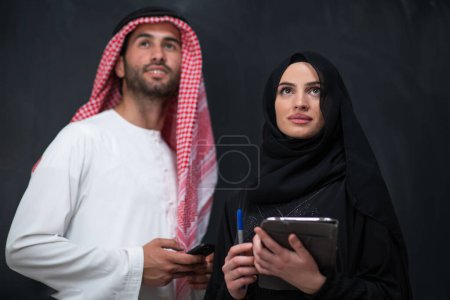 Téléchargez les photos : Young muslim business couple arabian man with woman in fashionable hijab dress using mobile phone and tablet computer in front of black chalkboard representing modern islam fashion technology. - en image libre de droit