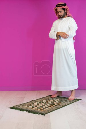Photo for Young arabian muslim man in traditional clothes praying on the carpet in front of pink wall before iftar dinner during a ramadan feast at home. - Royalty Free Image