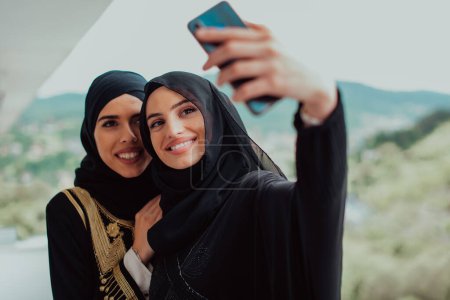 Foto de Jóvenes hermosas mujeres musulmanas en vestido de moda con hijab utilizando el teléfono móvil mientras toma foto selfie en el balcón que representa la tecnología moderna de la moda islam y el concepto kareem ramadán
. - Imagen libre de derechos