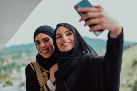Téléchargez les photos : Jeunes belles femmes musulmanes en robe à la mode avec hijab en utilisant le téléphone mobile tout en prenant des photos selfie sur le balcon représentant la technologie moderne de la mode islam et le concept de Ramadan Kareem
. - en image libre de droit