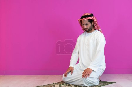 Foto de Joven musulmán árabe con ropa tradicional rezando en la alfombra frente a la pared rosa antes de la cena de iftar durante una fiesta de ramadán en casa. - Imagen libre de derechos
