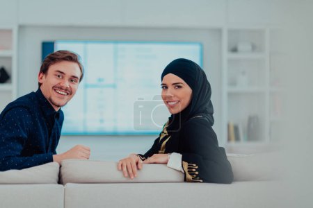 Téléchargez les photos : Jeune musulmane couple femme portant islamic hijab vêtements assis sur canapé regarder la télévision ensemble au cours du mois de Ramadan à la maison moderne
. - en image libre de droit