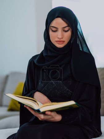 Photo for Young traditional muslim woman reading Quran on the sofa before iftar dinner during a ramadan feast at home. - Royalty Free Image