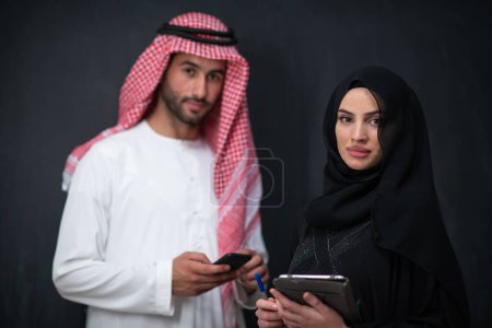 Photo for Young muslim business couple arabian man with woman in fashionable hijab dress using mobile phone and tablet computer in front of black chalkboard representing modern islam fashion technology. - Royalty Free Image