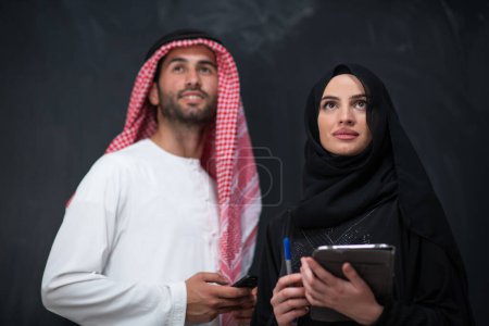Téléchargez les photos : Young muslim business couple arabian man with woman in fashionable hijab dress using mobile phone and tablet computer in front of black chalkboard representing modern islam fashion technology. - en image libre de droit