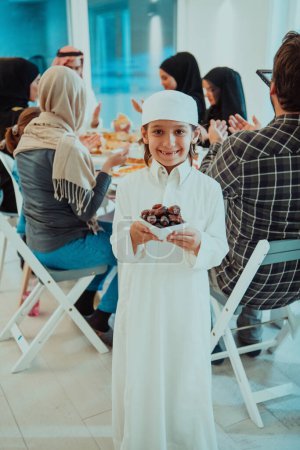 Foto de Eid Mubarak familia musulmana teniendo Iftar cena pequeño niño sosteniendo un plato lleno de dulces fechas para romper la fiesta. Comer comida tradicional durante el mes de fiesta del Ramadán en casa. La Comida Halal Islámica . - Imagen libre de derechos