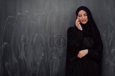 Photo for Young modern muslim business woman using smartphone wearing hijab clothes in front of black chalkboard. - Royalty Free Image