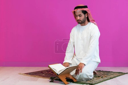 Téléchargez les photos : Jeune homme musulman arabe en vêtements traditionnels lisant le saint livre Coran sur le tapis de prière avant le dîner iftar lors d'une fête du ramadan à la maison
. - en image libre de droit