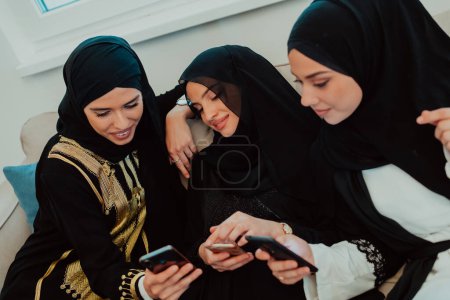 Téléchargez les photos : Group of young beautiful muslim women in fashionable dress with hijab using mobile phone while sitting on the sofa near window at home representing modern islam fashion technology and ramadan kareem. - en image libre de droit