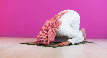 Téléchargez les photos : Jeune homme musulman arabe en vêtements traditionnels priant sur le tapis devant le mur rose avant le dîner iftar lors d'un festin de ramadan à la maison. - en image libre de droit