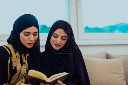 Foto de Two Young traditional Muslim women read Quran on the sofa before iftar dinner during a Ramadan feast at home. - Imagen libre de derechos