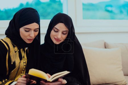Téléchargez les photos : Two Young traditional Muslim women read Quran on the sofa before iftar dinner during a Ramadan feast at home. - en image libre de droit