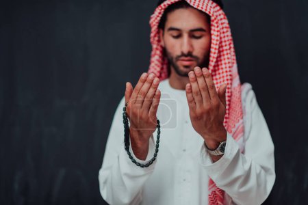 Photo for Arabian man in traditional clothes making traditional prayer to God, keeps hands in praying gesture in front of black chalkboard representing modern islam fashion and ramadan kareem concept. - Royalty Free Image
