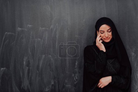 Téléchargez les photos : Jeune femme d'affaires musulmane moderne utilisant un smartphone portant des vêtements en hijab devant un tableau noir. - en image libre de droit