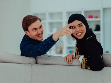 Téléchargez les photos : Jeune musulmane couple femme portant islamic hijab vêtements assis sur canapé regarder la télévision ensemble au cours du mois de Ramadan à la maison moderne
. - en image libre de droit
