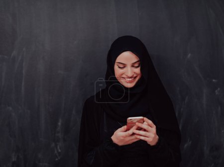 Photo for Young modern muslim business woman using smartphone wearing hijab clothes in front of black chalkboard. - Royalty Free Image