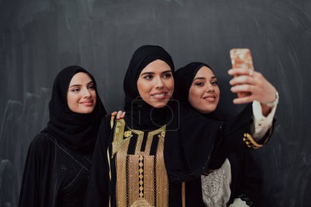 Photo for Group of young beautiful muslim women in fashionable dress with hijab using smartphone while taking selfie picture in front of black background. - Royalty Free Image