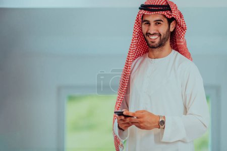 Foto de Young modern Muslim Arabian businessmen wearing traditional clothes while using smartphones at home - Imagen libre de derechos