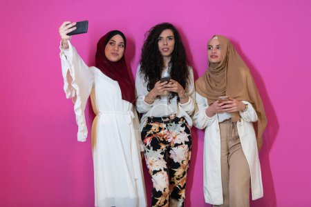 Foto de Group of beautiful muslim women two of them in fashionable dress with hijab using mobile phone while taking selfie picture isolated on pink background representing modern islam fashion technology. - Imagen libre de derechos