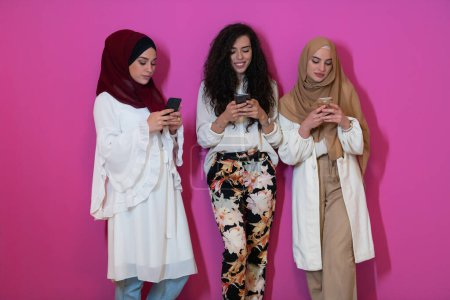 Téléchargez les photos : Groupe de belles femmes musulmanes deux d'entre eux en robe à la mode avec hijab en utilisant un téléphone mobile isolé sur fond rose représentant la technologie moderne de la mode islam et le concept de Ramadan Kareem
. - en image libre de droit
