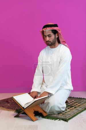 Foto de Joven musulmán árabe vestido con ropa tradicional leyendo el libro sagrado Corán en la alfombra de oración antes de la cena de iftar durante una fiesta de ramadán en casa
. - Imagen libre de derechos