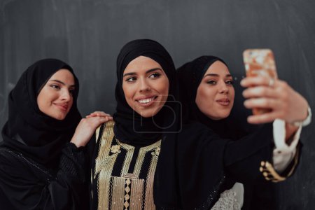 Téléchargez les photos : Group of young beautiful muslim women in fashionable dress with hijab using smartphone while taking selfie picture in front of black background. - en image libre de droit