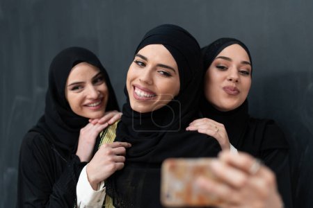 Téléchargez les photos : Group of young beautiful muslim women in fashionable dress with hijab using smartphone while taking selfie picture in front of black background. - en image libre de droit