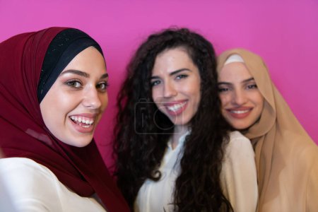 Téléchargez les photos : Group of beautiful muslim women two of them in fashionable dress with hijab using mobile phone while taking selfie picture isolated on pink background representing modern islam fashion technology. - en image libre de droit