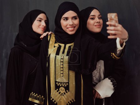 Photo for Group of young beautiful muslim women in fashionable dress with hijab using smartphone while taking selfie picture in front of black background. - Royalty Free Image