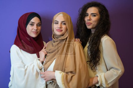 Téléchargez les photos : Portrait de groupe de belles femmes musulmanes deux d'entre eux en robe à la mode avec hijab isolé sur fond violet représentant la mode islam moderne et concept de Ramadan Kareem
. - en image libre de droit
