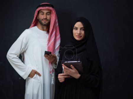 Téléchargez les photos : Young muslim business couple arabian man with woman in fashionable hijab dress using mobile phone and tablet computer in front of black chalkboard representing modern islam fashion technology . - en image libre de droit