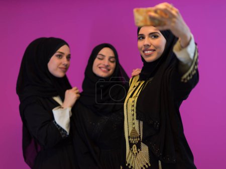 Téléchargez les photos : Group of beautiful muslim women two of them in fashionable dress with hijab using mobile phone while taking selfie picture isolated on pink background representing modern islam fashion technology. - en image libre de droit