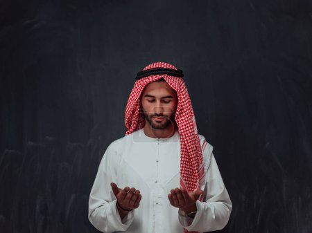 Téléchargez les photos : Arabian man in traditional clothes making traditional prayer to God, keeps hands in praying gesture in front of black chalkboard representing modern islam fashion and ramadan kareem concept. - en image libre de droit