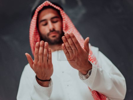 Foto de Arabian man in traditional clothes making traditional prayer to God, keeps hands in praying gesture in front of black chalkboard representing modern islam fashion and ramadan kareem concept. - Imagen libre de derechos