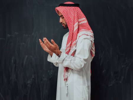 Foto de Arabian man in traditional clothes making traditional prayer to God, keeps hands in praying gesture in front of black chalkboard representing modern islam fashion and ramadan kareem concept. - Imagen libre de derechos