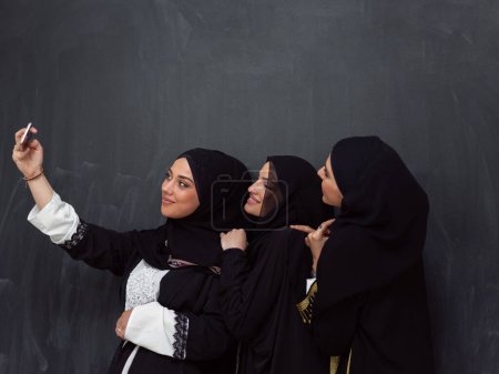 Foto de Group of young beautiful muslim women in fashionable dress with hijab using smartphone while taking selfie picture in front of black background. - Imagen libre de derechos