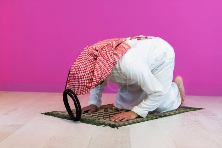 Téléchargez les photos : Jeune homme musulman arabe en vêtements traditionnels priant sur le tapis devant le mur rose avant le dîner iftar lors d'un festin de ramadan à la maison. - en image libre de droit