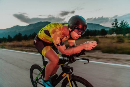 Photo for Night drive. Full length portrait of an active triathlete in sportswear and with a protective helmet riding a bicycle in night time.. Selective focus. - Royalty Free Image