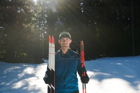 Foto de Retrato apuesto atleta masculino con esquís de fondo en las manos y gafas, entrenamiento en el bosque nevado. Concepto de estilo de vida saludable invierno - Imagen libre de derechos