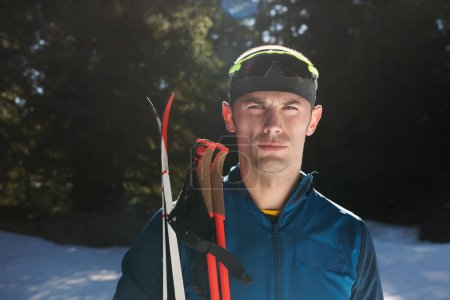 Téléchargez les photos : Portrait bel athlète masculin avec des skis de fond dans les mains et les lunettes, l'entraînement dans la forêt enneigée. Concept de mode de vie sain hiver - en image libre de droit