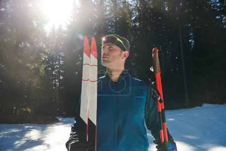 Téléchargez les photos : Portrait bel athlète masculin avec des skis de fond dans les mains et les lunettes, l'entraînement dans la forêt enneigée. Concept de mode de vie sain hiver - en image libre de droit