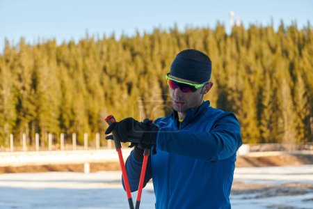 Téléchargez les photos : Athlète masculin séduisant avec des skis de fond, respirant fraîchement et faisant une pause après un entraînement difficile dans une forêt enneigée. Je vérifie les montres intelligentes. Mode de vie sain hiver. - en image libre de droit