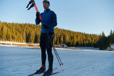 Téléchargez les photos : Beau athlète masculin avec des skis de fond préparant l'équipement pour l'entraînement dans une forêt enneigée. Je vérifie les montres intelligentes. Mode de vie sain hiver - en image libre de droit