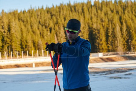 Téléchargez les photos : Athlète masculin séduisant avec des skis de fond, respirant fraîchement et faisant une pause après un entraînement difficile dans une forêt enneigée. Je vérifie les montres intelligentes. Mode de vie sain hiver. - en image libre de droit