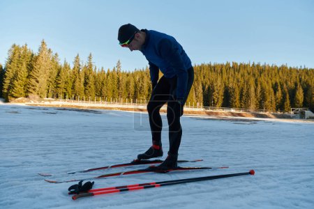 Téléchargez les photos : Handsome male athlete with cross country skis preparing equipment for training in a snowy forest. Checking smartwatch. Healthy winter lifestyle - en image libre de droit