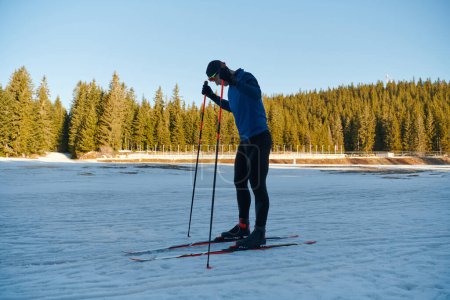 Téléchargez les photos : Beau athlète masculin avec des skis de fond préparant l'équipement pour l'entraînement dans une forêt enneigée. Je vérifie les montres intelligentes. Mode de vie sain hiver - en image libre de droit