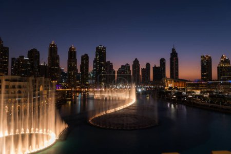 Photo for Dubai singing fountains at night lake view between skyscrapers. City skyline in dusk modern architecture in UAE capital downtown. High quality 4k footage - Royalty Free Image