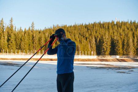 Photo for Handsome male athlete with cross country skis preparing equipment for training in a snowy forest. Checking smartwatch. Healthy winter lifestyle - Royalty Free Image