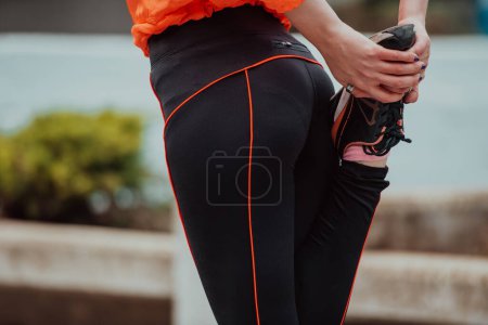 Téléchargez les photos : Fit attractive woman in sportswear stretching one leg before jogging on the footpath outdoor in summer among greenery. Workout, sport, activity, fitness, vacation and training conept - en image libre de droit