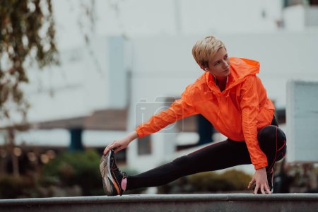 Foto de Fit attractive woman in sportswear stretching one leg before jogging on the footpath outdoor in summer among greenery. Workout, sport, activity, fitness, vacation and training conept - Imagen libre de derechos
